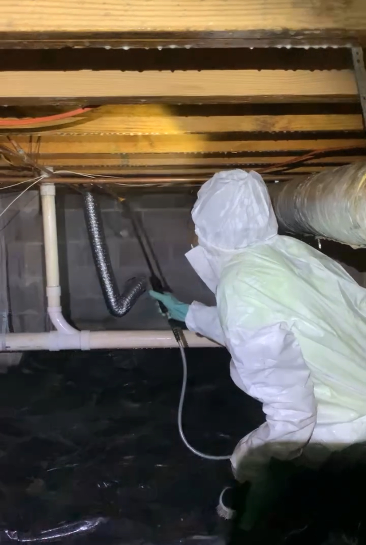 A person in protective gear sprays a substance onto pipes and beams in a dimly lit crawl space.