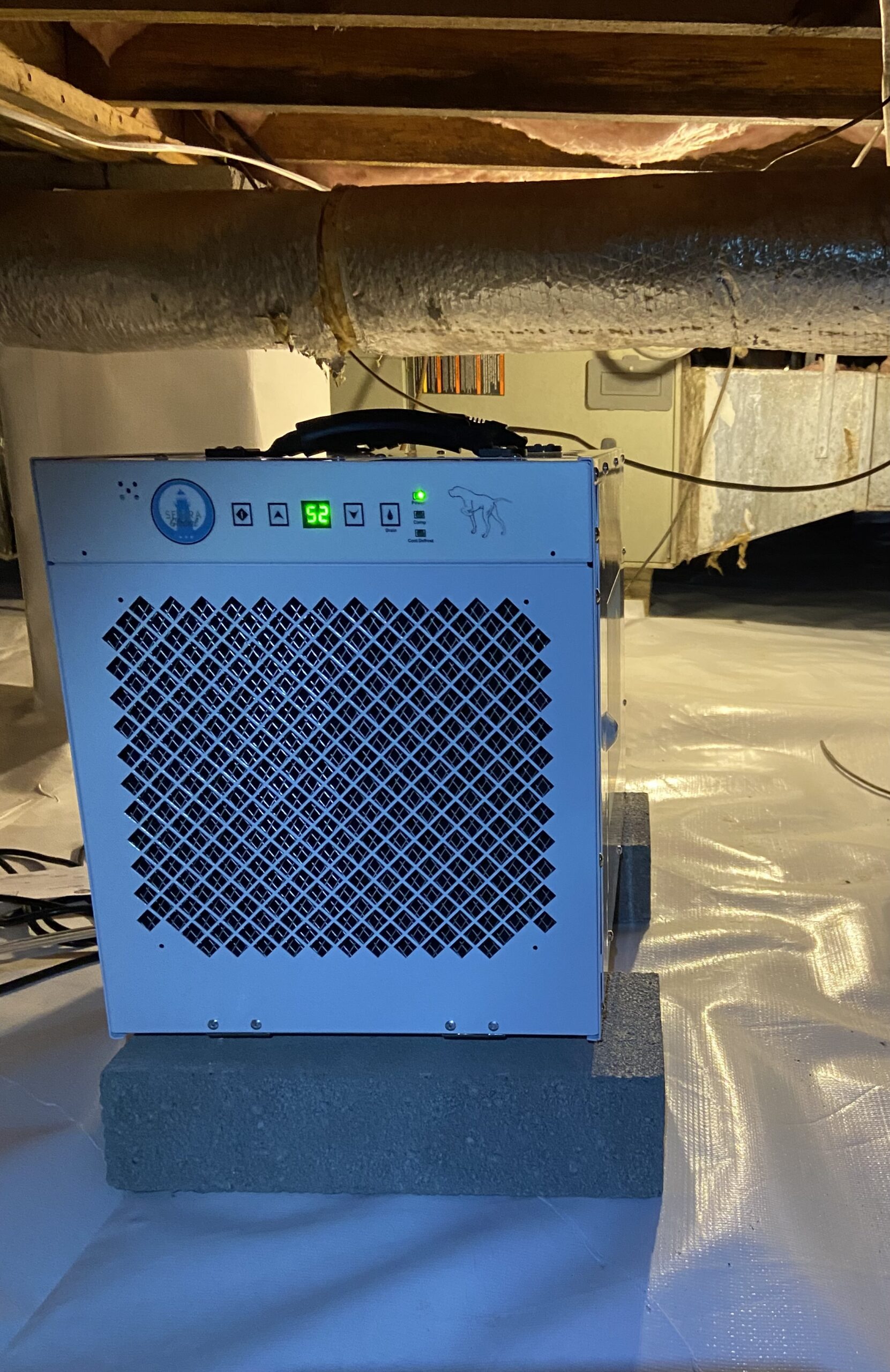 A dehumidifier sits on cinder blocks in a crawl space with white plastic vapor barrier and exposed insulated pipes overhead.