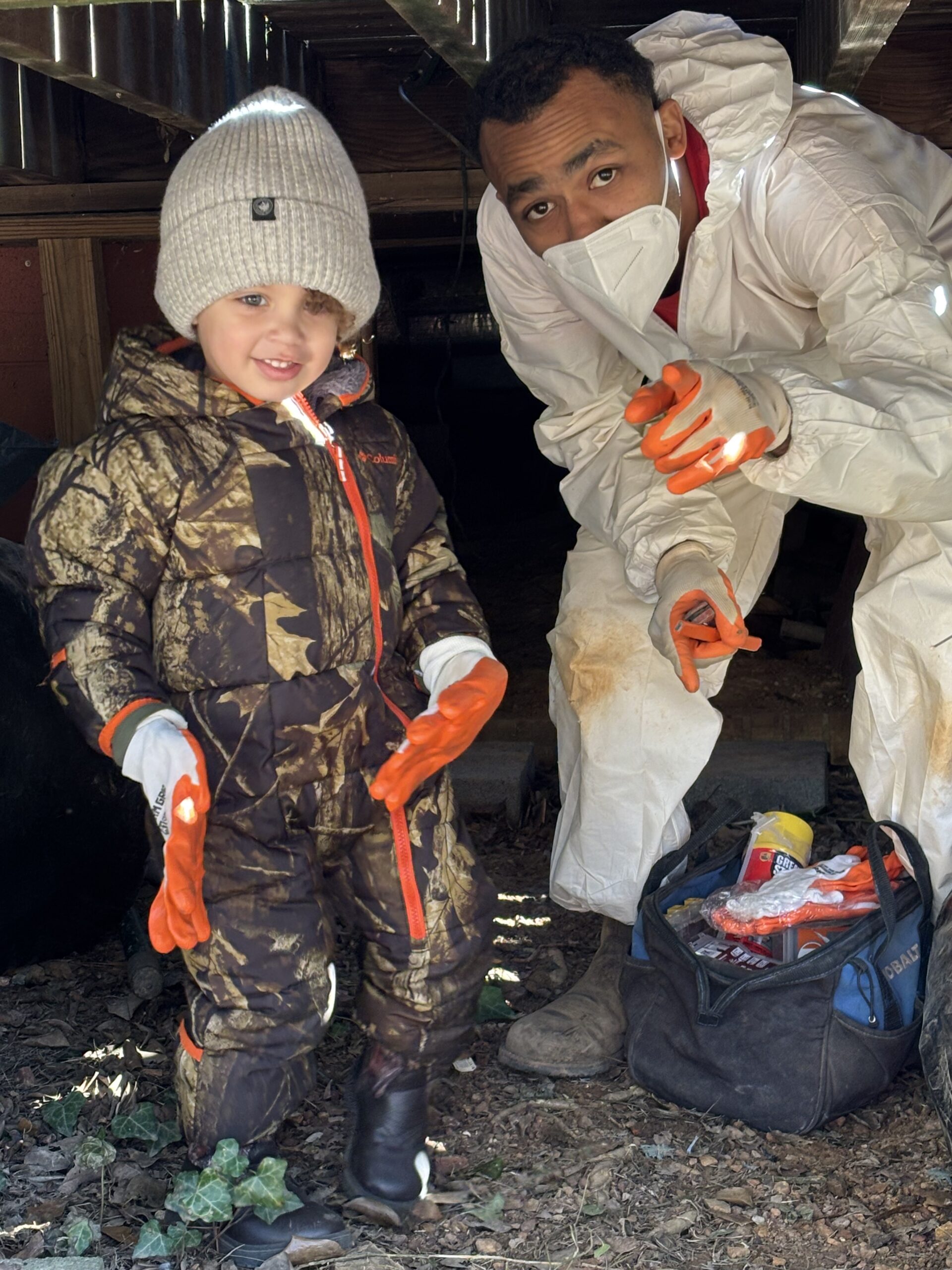 A young child in camouflage winter gear and an adult in a white protective suit and mask, both wearing gloves, stand under a structure—possibly inspecting it for a waterproofing service—next to a bag of supplies.