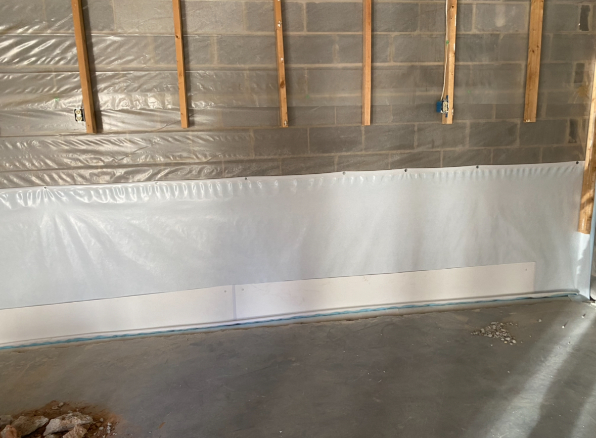Concrete basement wall with plastic vapor barrier partially installed, wooden studs exposed, and unfinished floor visible.