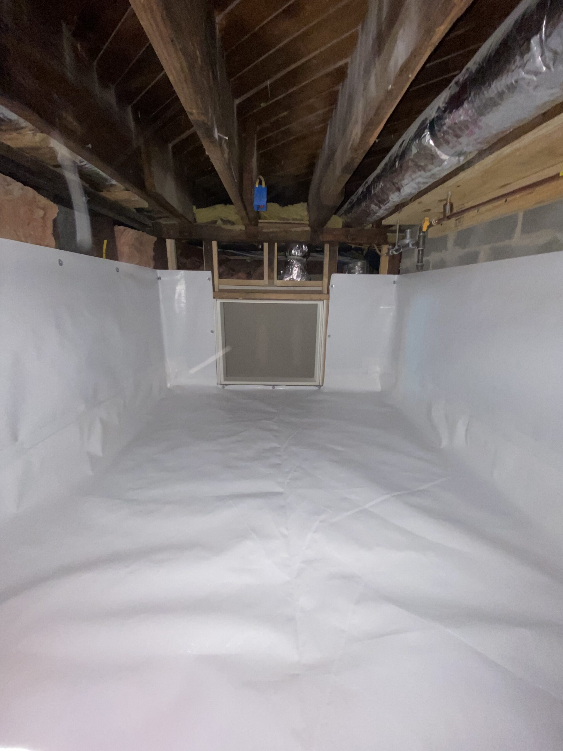 A crawl space with a white vapor barrier lining the floor and walls, exposed wooden beams above, ductwork, and a vent or window at the far end.