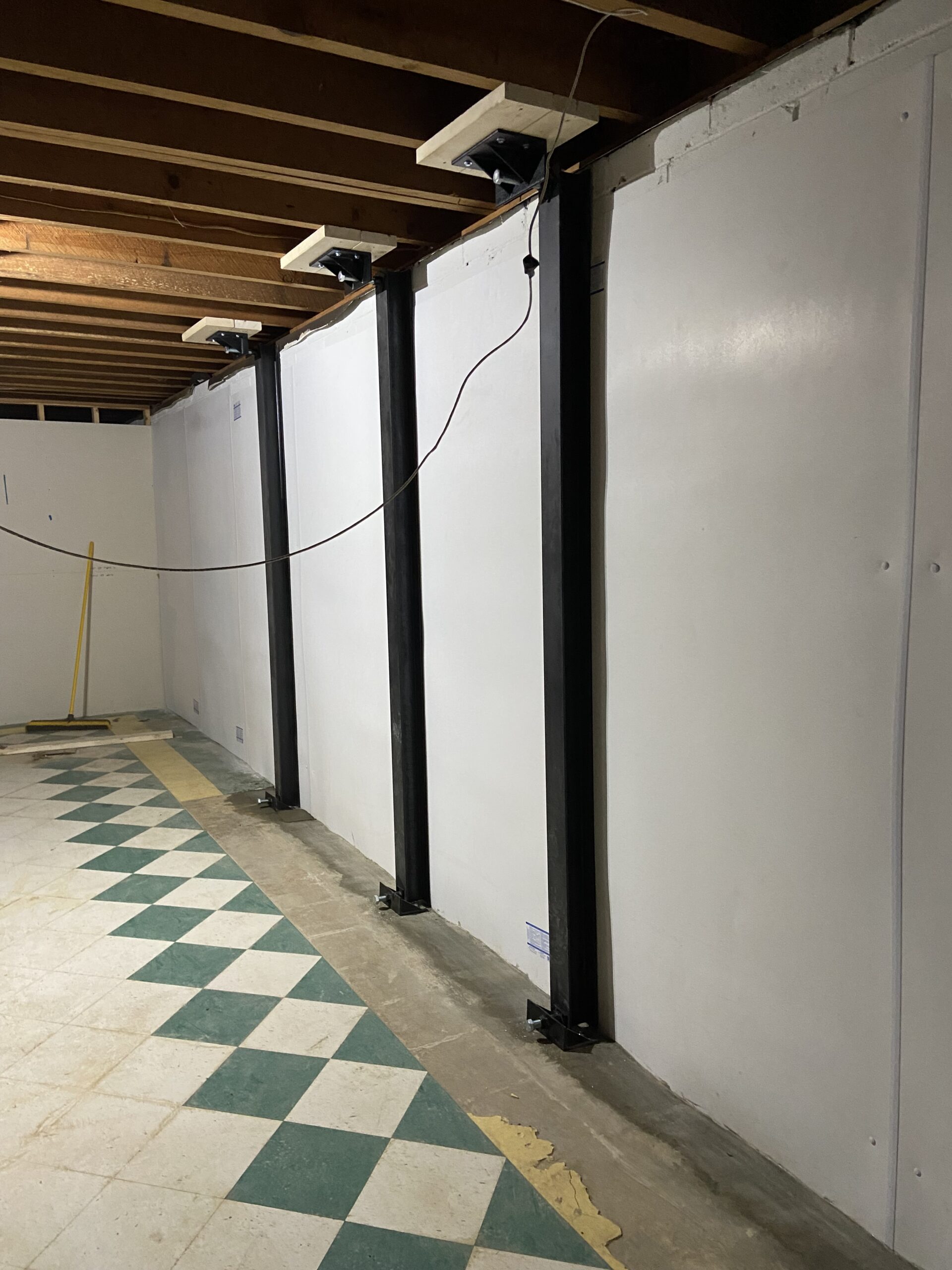 Basement with exposed ceiling beams, tiled floor, and three vertical steel support beams installed against a white wall. A broom leans against the far wall.