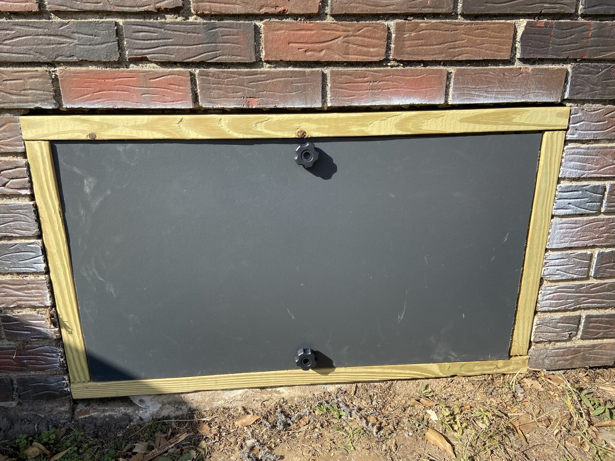 A rectangular crawl space cover with a wooden frame and two black fasteners, installed in a brick wall near the ground.