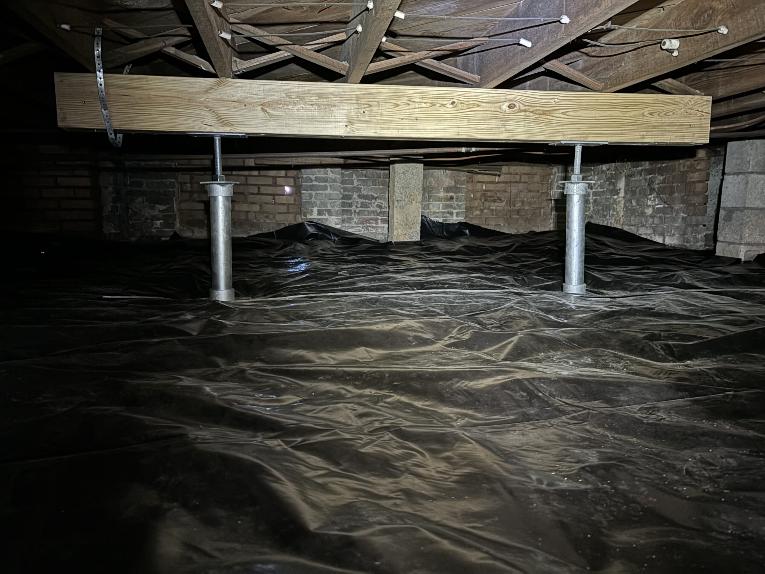 A wooden support beam is held up by two metal jack posts in a crawl space with a plastic vapor barrier on the ground.