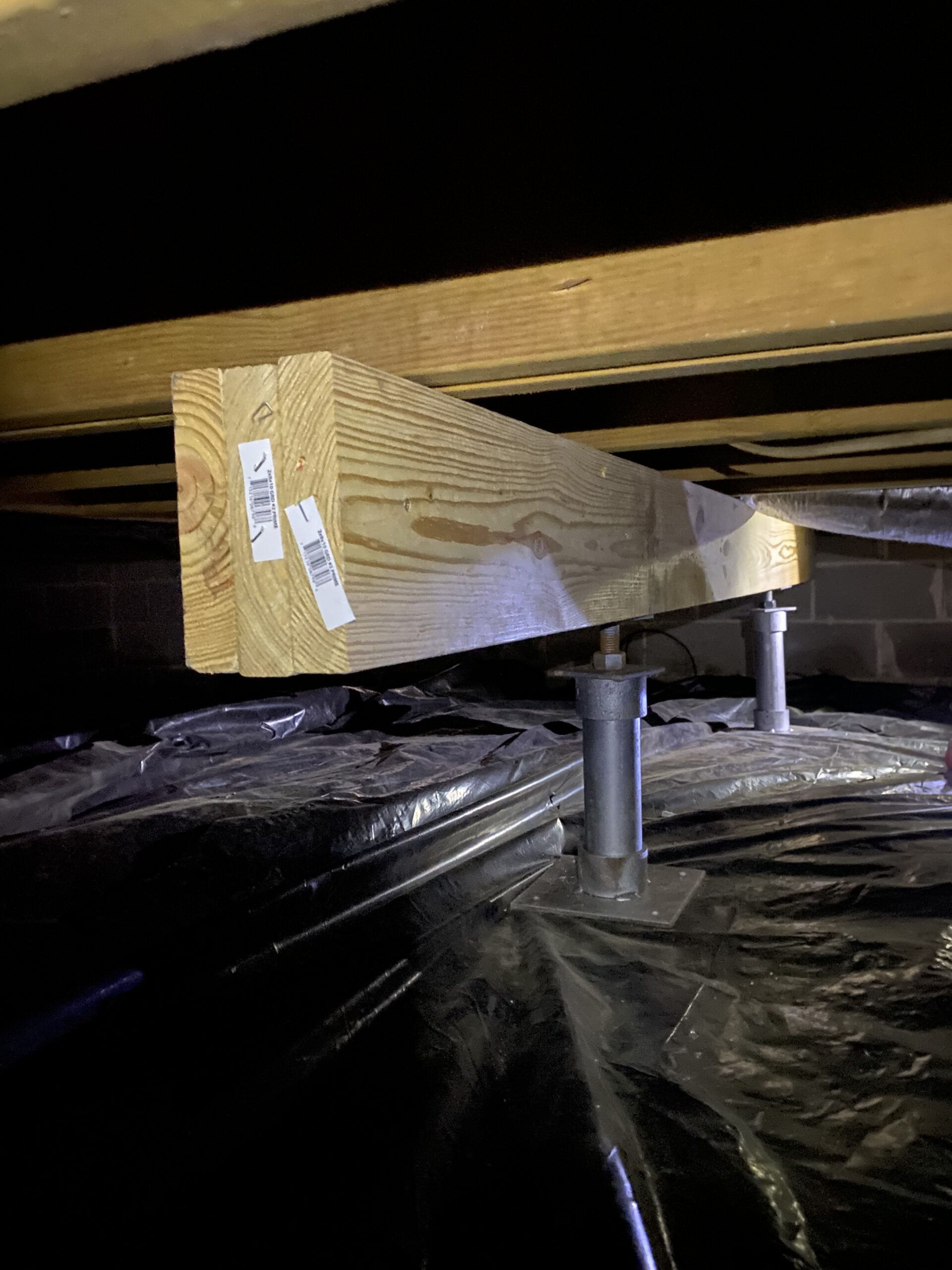 A wooden support beam rests on metal adjustable jacks above a black plastic vapor barrier in a crawl space.