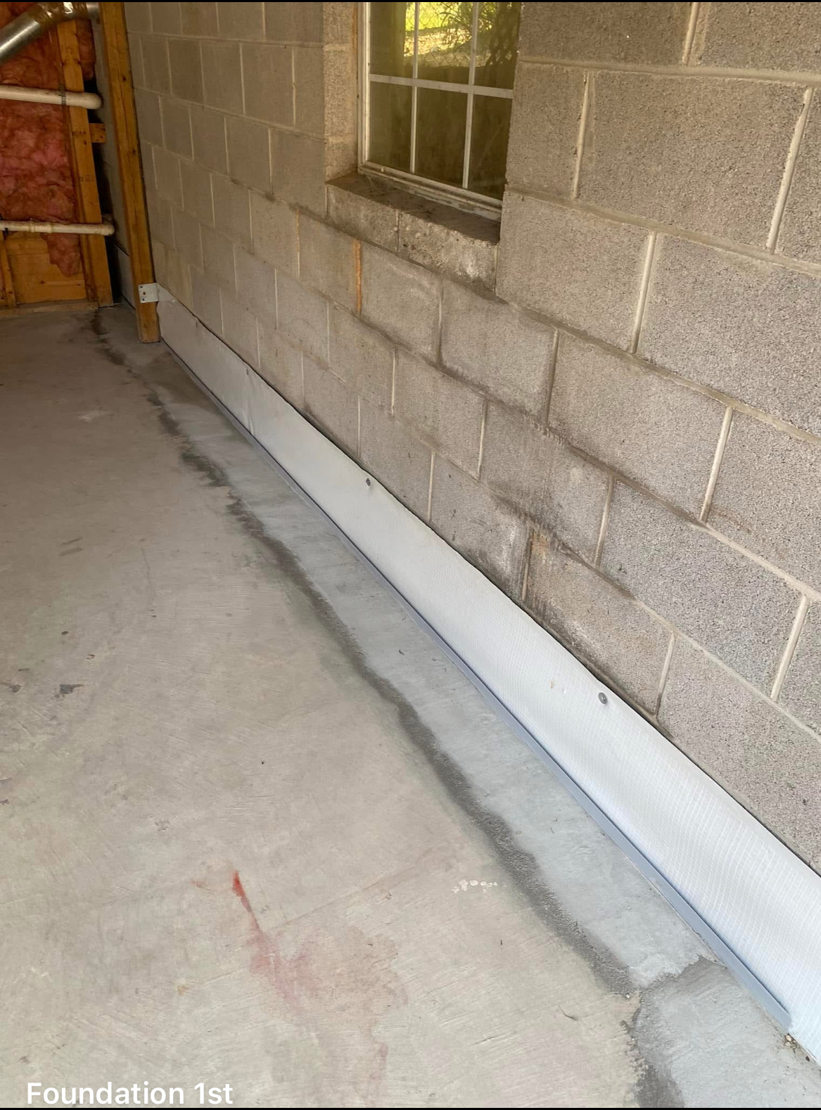 Concrete basement wall with a drainage system installed along the base, featuring a white barrier against the wall and a visible concrete seam on the floor.