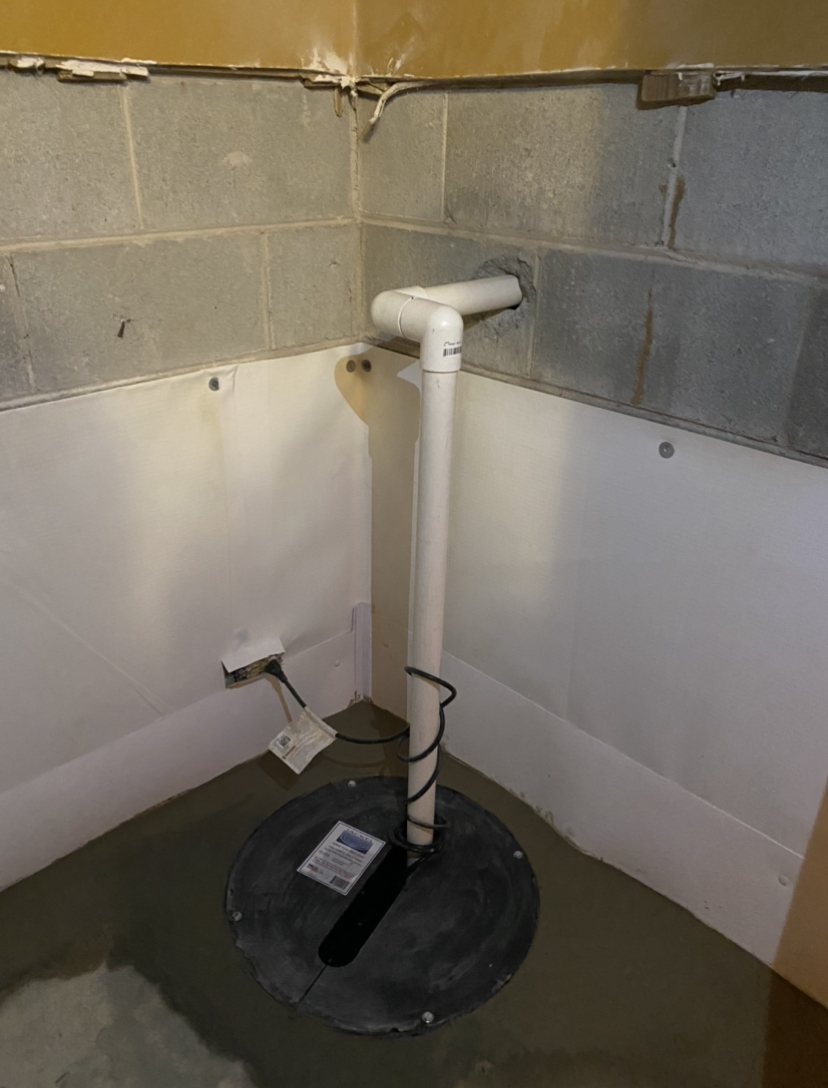 Sump pump system installed in a basement corner, with PVC discharge pipe running into a cinder block wall and a power cord plugged into an outlet.